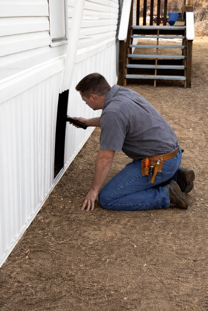 Technician inspecting underneath a mobile home through skirting access panel during underhome inspection in Florida